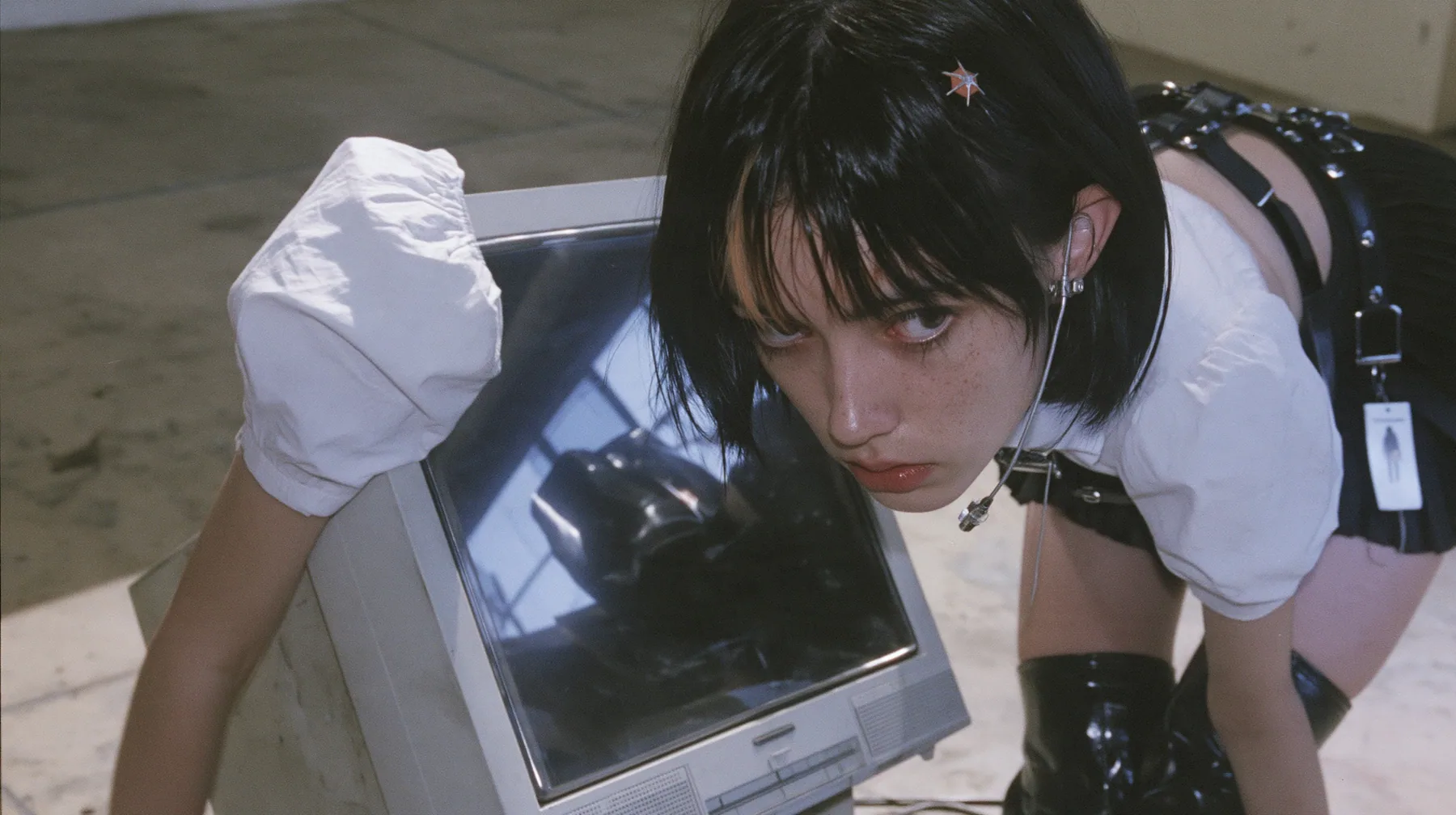 an old boxy monitor lying on a concrete floor with a black screen, the face of a young Caucasian American woman with pale skin and light freckles, a glossy black blunt jaw-length bob with heavy bangs and one…