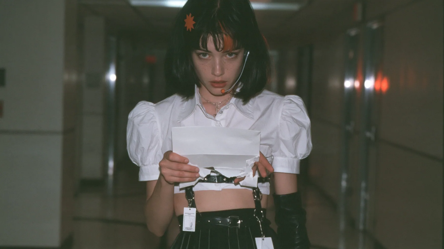 a young Caucasian American woman with pale skin and light freckles, a glossy black blunt jaw-length bob with heavy bangs and one clay-orange streak through the bangs, a small flat clay-orange eight-pointed star hair…