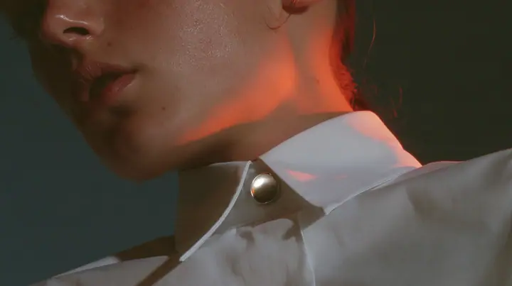 extreme close-up of a small round blank enamel pin on the collar of a white cropped shirt, jaw and the orange streak at the top edge of frame, pre-dawn slate grey, one small ember-orange #db5c34 light as the only…
