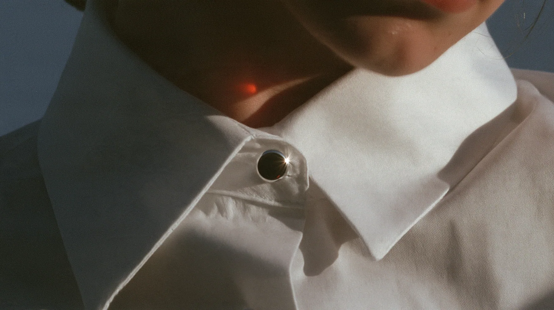 extreme close-up of a small round blank enamel pin on the collar of a white cropped shirt, jaw and the orange streak at the top edge of frame, pre-dawn slate grey, one small ember-orange #db5c34 light as the only…