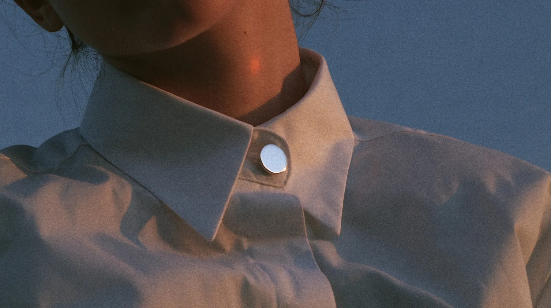 extreme close-up of a small round blank enamel pin on the collar of a white cropped shirt, jaw and the orange streak at the top edge of frame, pre-dawn slate grey, one small ember-orange #db5c34 light as the only…