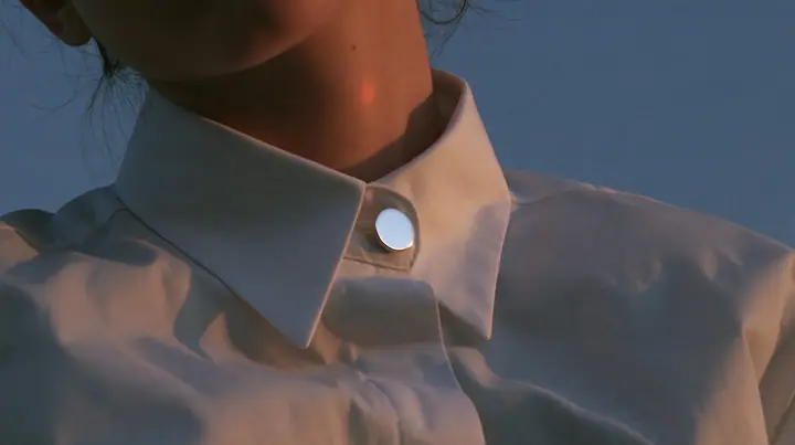 extreme close-up of a small round blank enamel pin on the collar of a white cropped shirt, jaw and the orange streak at the top edge of frame, pre-dawn slate grey, one small ember-orange #db5c34 light as the only…
