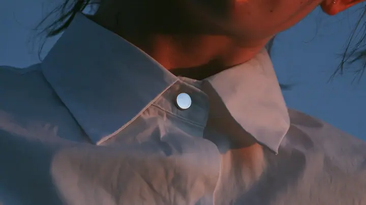 extreme close-up of a small round blank enamel pin on the collar of a white cropped shirt, jaw and the orange streak at the top edge of frame, pre-dawn slate grey, one small ember-orange #db5c34 light as the only…