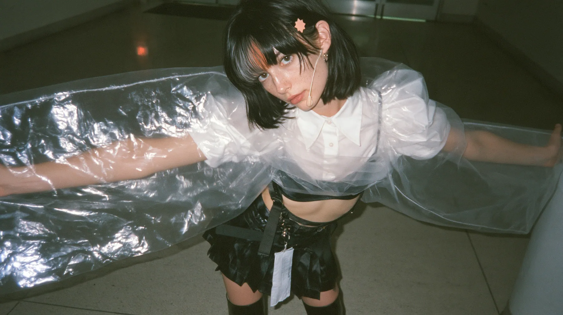 a young Caucasian American woman with pale skin and light freckles, a glossy black blunt jaw-length bob with heavy bangs and one clay-orange streak through the bangs, a small flat clay-orange eight-pointed star hair…