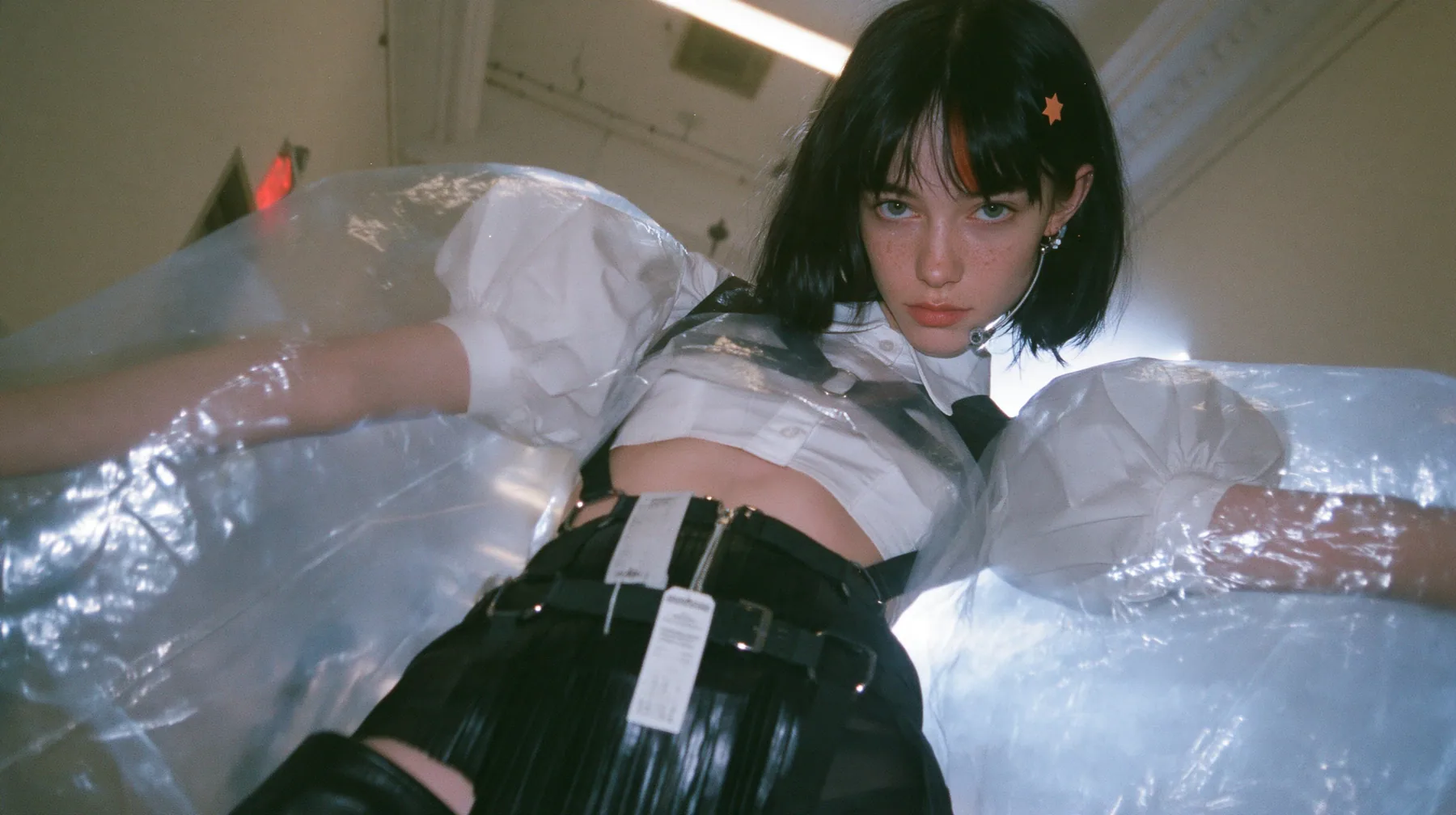 a young Caucasian American woman with pale skin and light freckles, a glossy black blunt jaw-length bob with heavy bangs and one clay-orange streak through the bangs, a small flat clay-orange eight-pointed star hair…