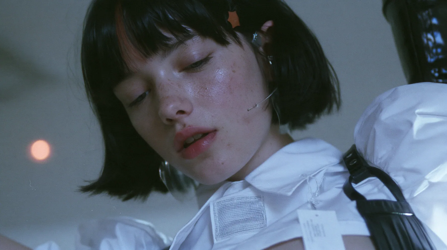 extreme close-up of a blank woven label stitched inside the collar of a white shirt, jaw of a young Caucasian American woman with pale skin and light freckles, a glossy black blunt jaw-length bob with heavy bangs and…