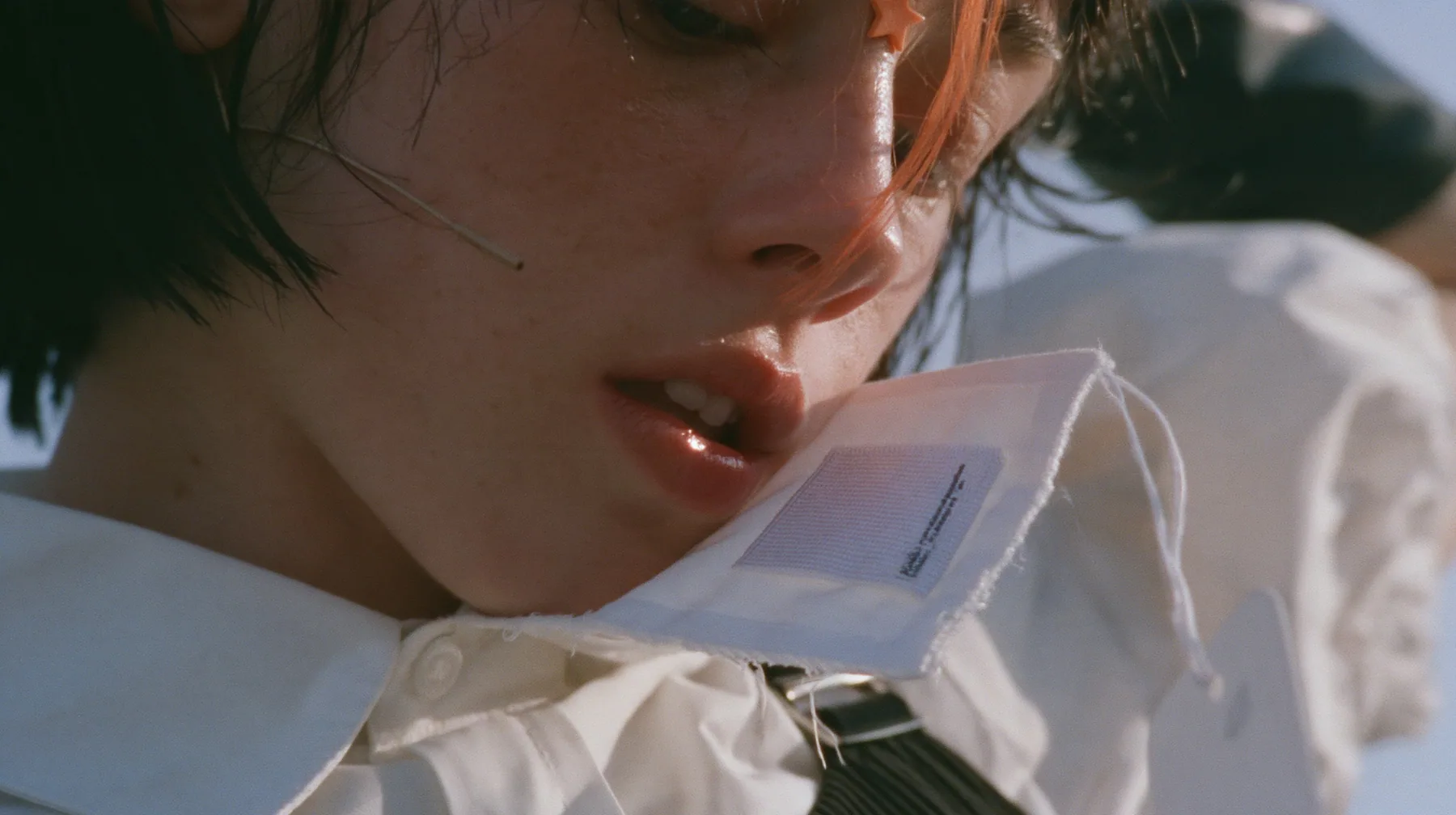 extreme close-up of a blank woven label stitched inside the collar of a white shirt, jaw of a young Caucasian American woman with pale skin and light freckles, a glossy black blunt jaw-length bob with heavy bangs and…