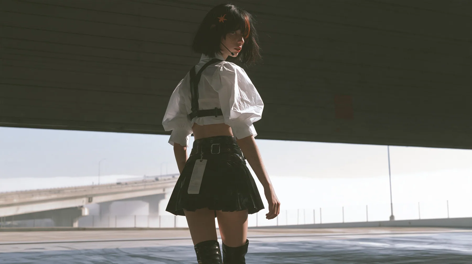 a huge rolling loading door rising at dawn, pale light flooding a dark hall, beyond it an elevated freeway above a sea of fog, a young Caucasian American woman with pale skin and light freckles, a glossy black blunt…