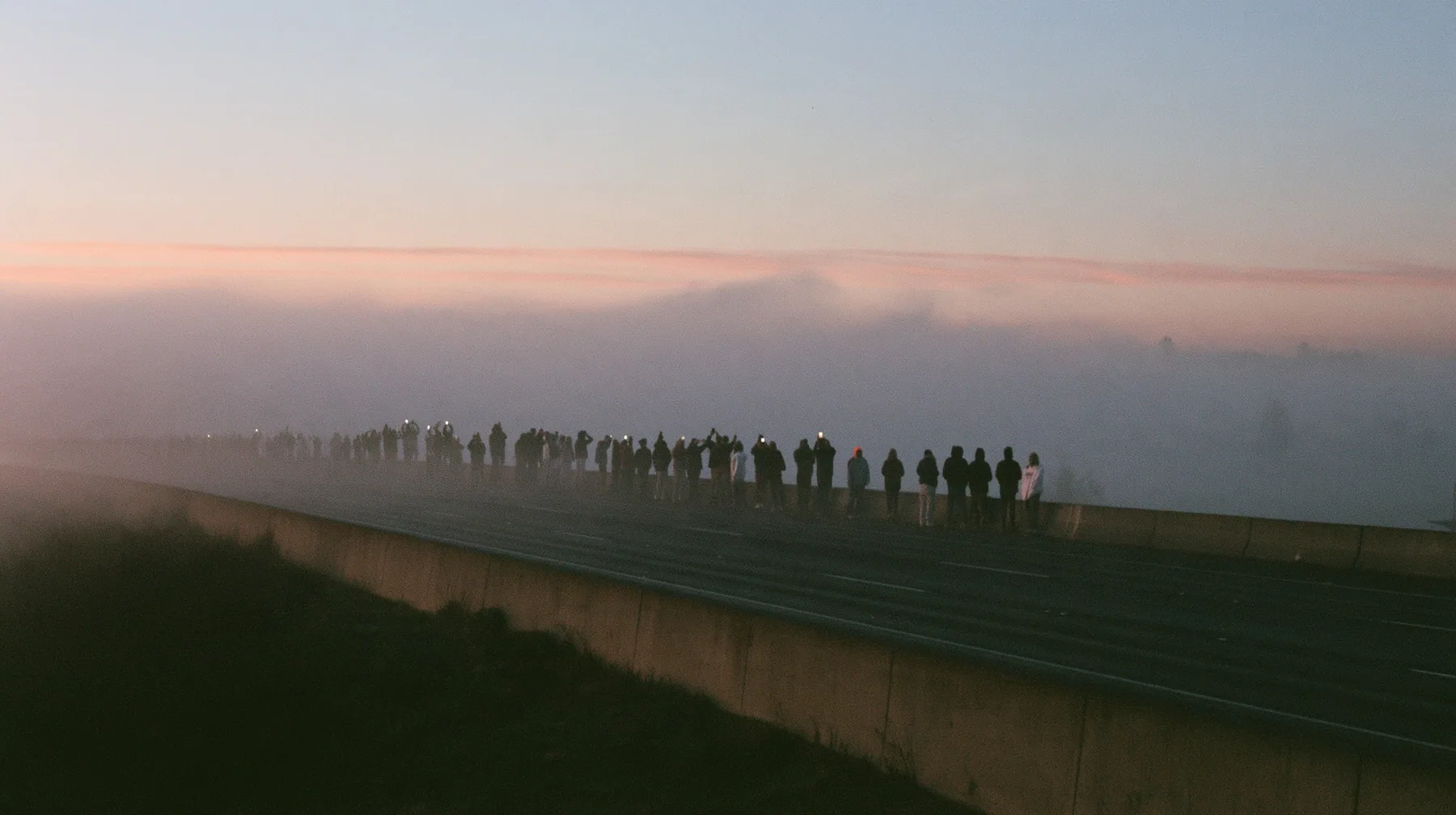 a line of spectators along a freeway barrier at dawn holding phones up, fog below, pale dawn grey-white, soft even light, a single clay-orange #D97757 accent, fine grain, calm, no text, no letters, no logos