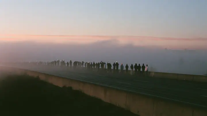 a line of spectators along a freeway barrier at dawn holding phones up, fog below, pale dawn grey-white, soft even light, a single clay-orange #D97757 accent, fine grain, calm, no text, no letters, no logos