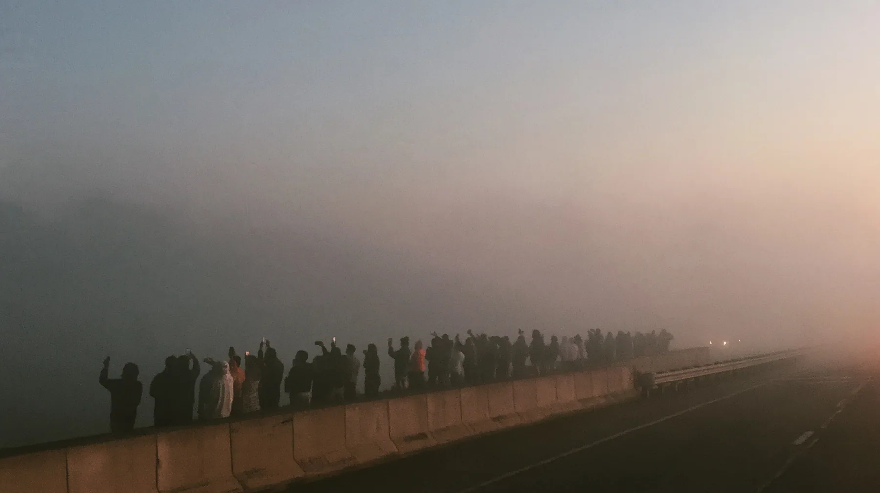 a line of spectators along a freeway barrier at dawn holding phones up, fog below, pale dawn grey-white, soft even light, a single clay-orange #D97757 accent, fine grain, calm, no text, no letters, no logos