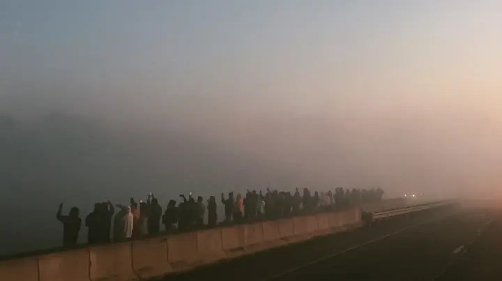 a line of spectators along a freeway barrier at dawn holding phones up, fog below, pale dawn grey-white, soft even light, a single clay-orange #D97757 accent, fine grain, calm, no text, no letters, no logos
