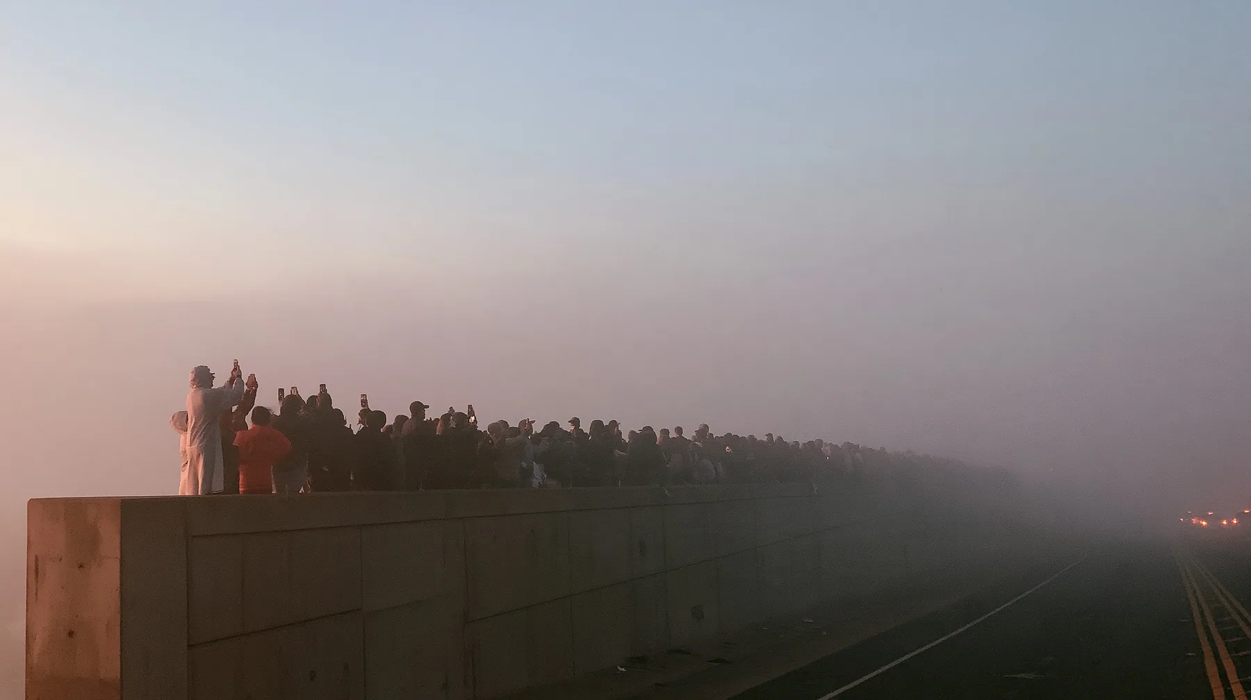 a line of spectators along a freeway barrier at dawn holding phones up, fog below, pale dawn grey-white, soft even light, a single clay-orange #D97757 accent, fine grain, calm, no text, no letters, no logos