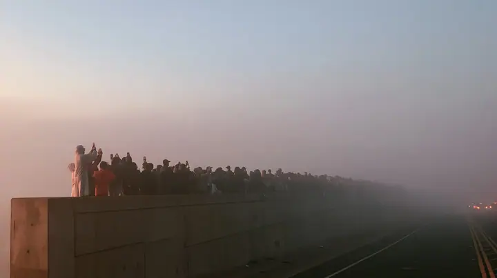 a line of spectators along a freeway barrier at dawn holding phones up, fog below, pale dawn grey-white, soft even light, a single clay-orange #D97757 accent, fine grain, calm, no text, no letters, no logos