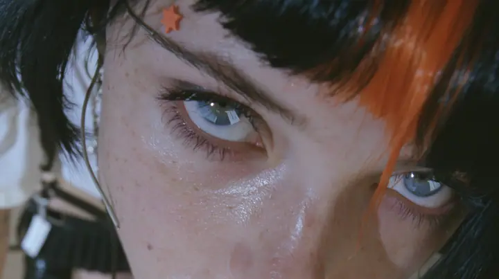extreme close-up of an eye of a young Caucasian American woman with pale skin and light freckles, a glossy black blunt jaw-length bob with heavy bangs and one clay-orange streak through the bangs, a small flat…