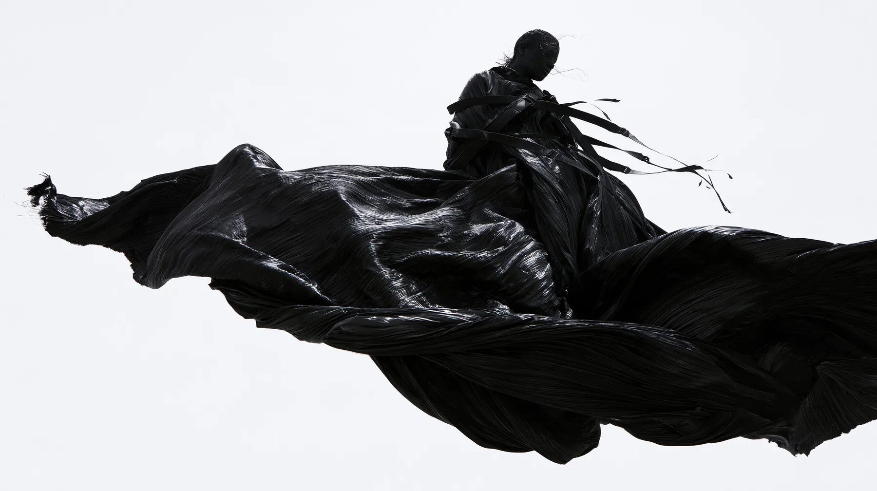 a huge black pleated fabric whipping in wind against a white void, harness straps flying, high-key white void, pure white paper light, hard clean black shadows, monochrome, no text, no letters, no logos