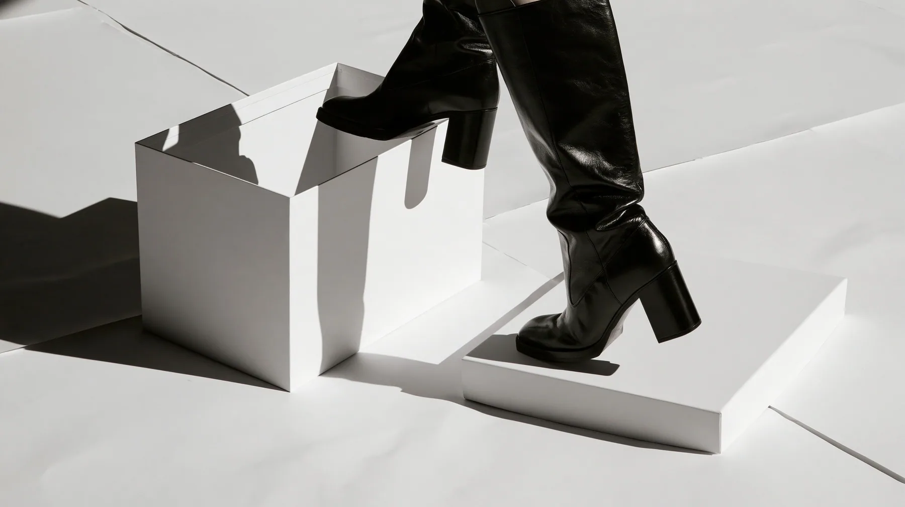 an empty white shoebox with its lid off on a white paper floor, one black knee boot stepping out of it, high-key white void, pure white paper light, hard clean black shadows, monochrome, no text, no letters, no logos