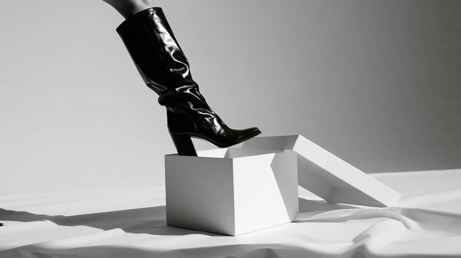 an empty white shoebox with its lid off on a white paper floor, one black knee boot stepping out of it, high-key white void, pure white paper light, hard clean black shadows, monochrome, no text, no letters, no logos