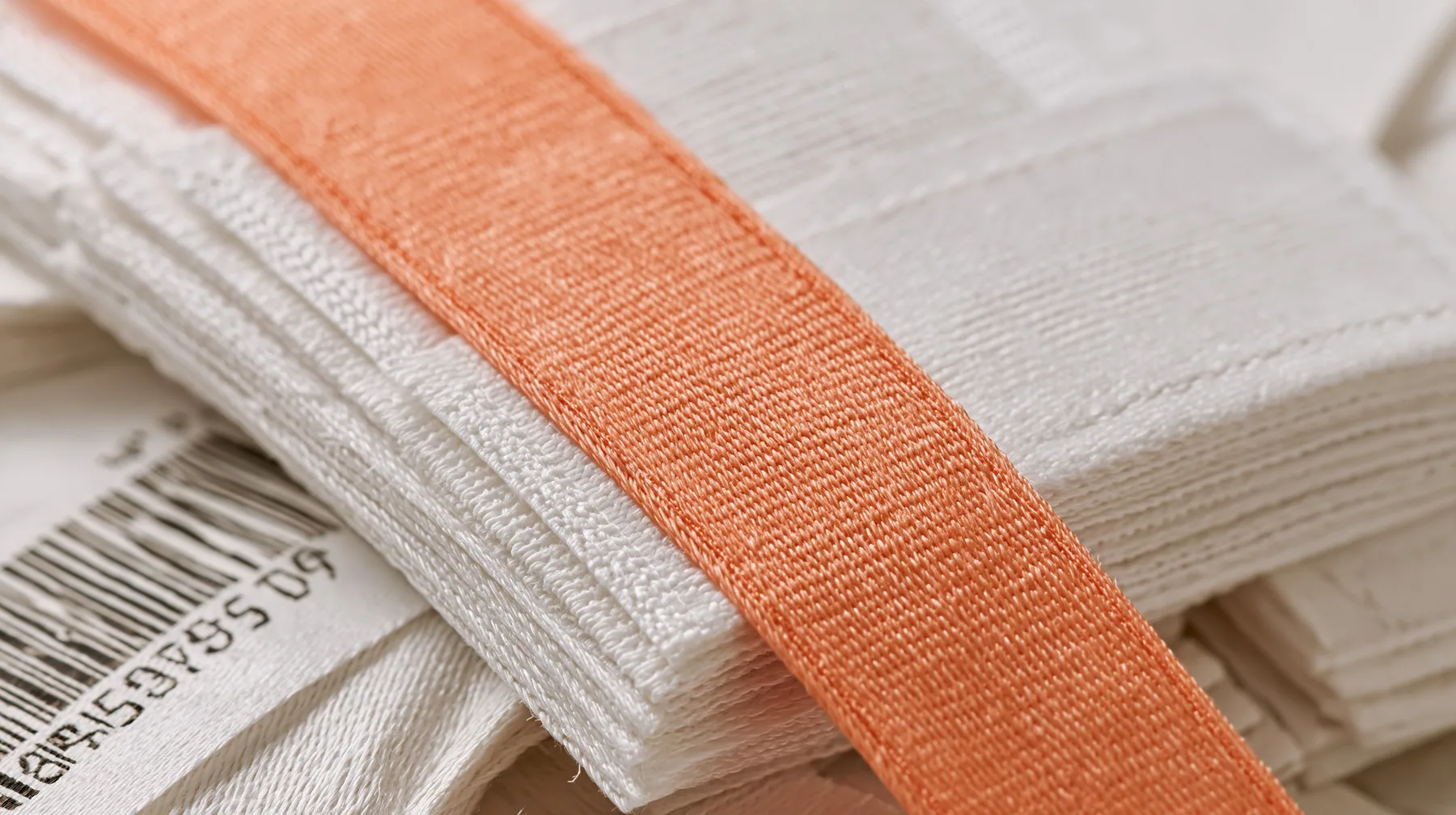 macro of a stack of white woven garment labels and one clay-orange label, barcodes, thread texture, high-key white paper, one clay-orange #D97757 accent as the only colour, soft shadowless light, no text, no letters,…