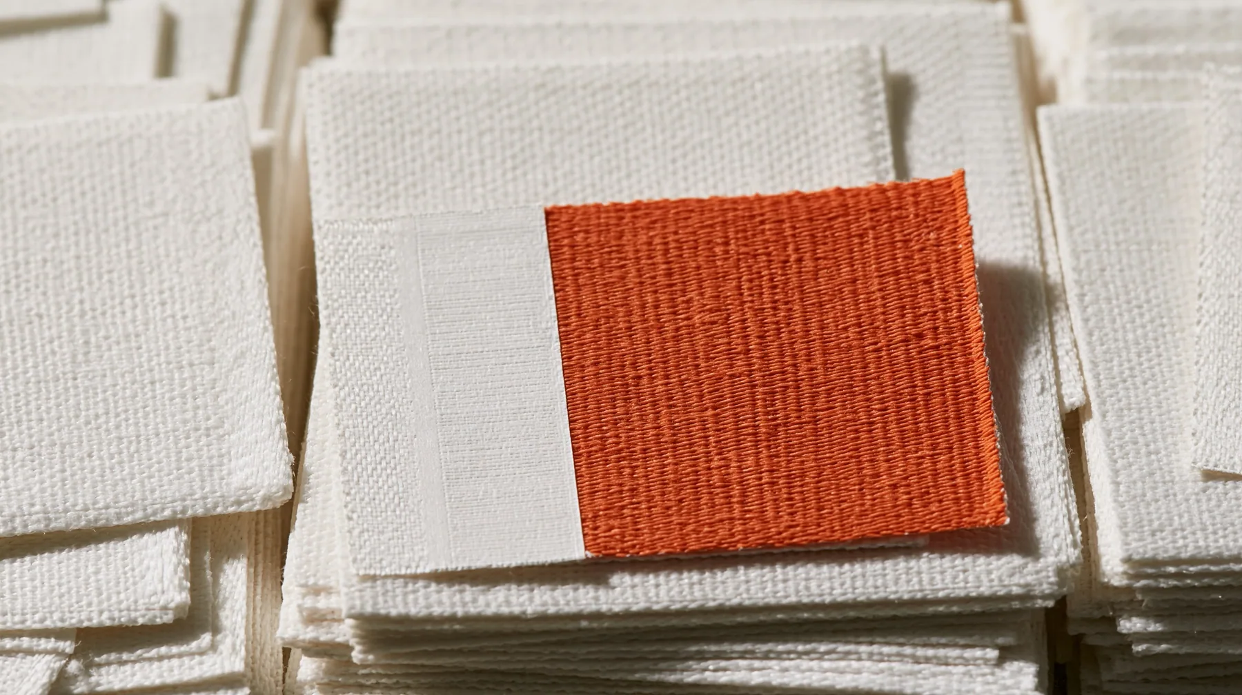 macro of a stack of white woven garment labels and one clay-orange label, barcodes, thread texture, high-key white paper, one clay-orange #D97757 accent as the only colour, soft shadowless light, no text, no letters,…