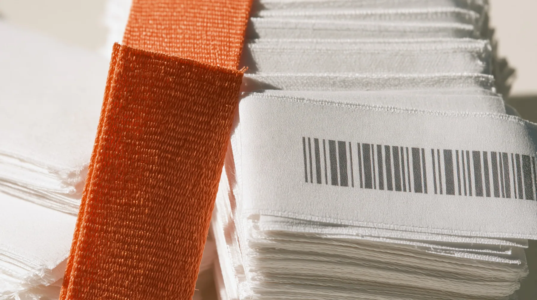 macro of a stack of white woven garment labels and one clay-orange label, barcodes, thread texture, high-key white paper, one clay-orange #D97757 accent as the only colour, soft shadowless light, no text, no letters,…