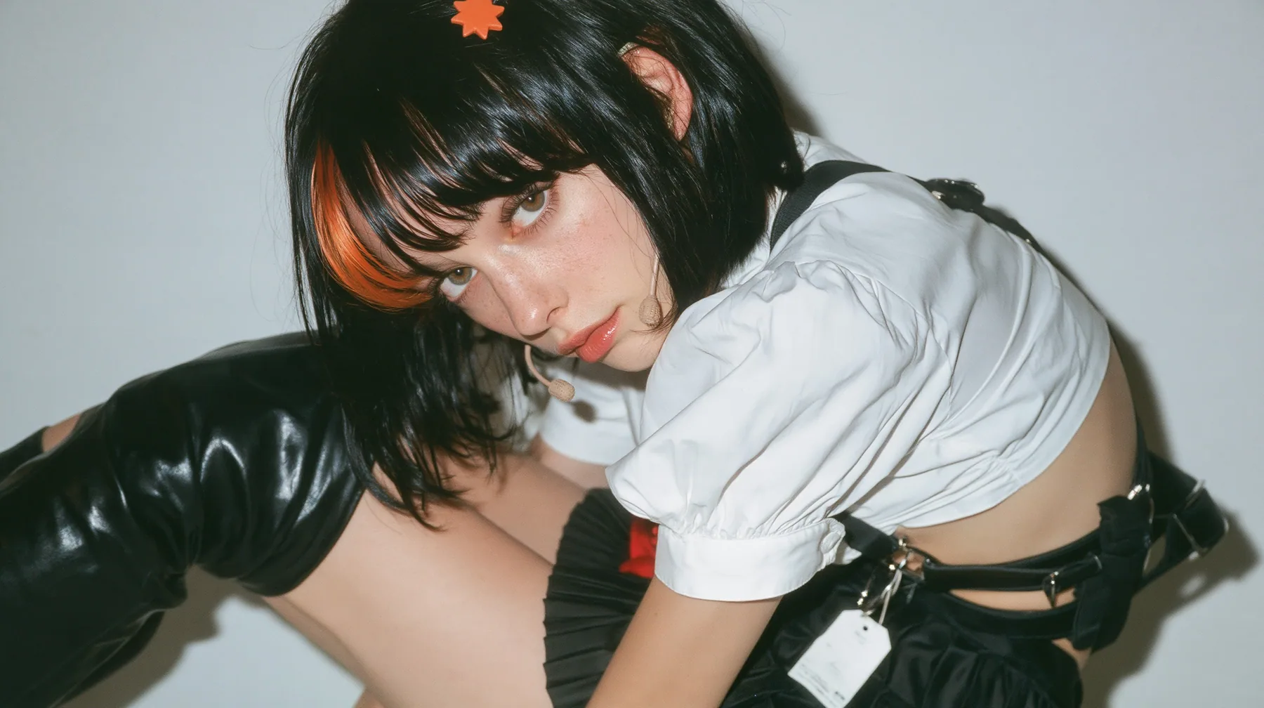 the hips of a young Caucasian American woman with pale skin and light freckles, a glossy black blunt jaw-length bob with heavy bangs and one clay-orange streak through the bangs, a small flat clay-orange…