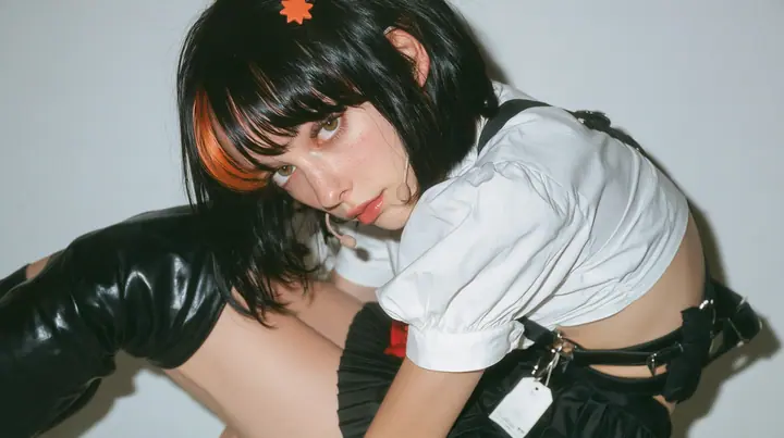 the hips of a young Caucasian American woman with pale skin and light freckles, a glossy black blunt jaw-length bob with heavy bangs and one clay-orange streak through the bangs, a small flat clay-orange…