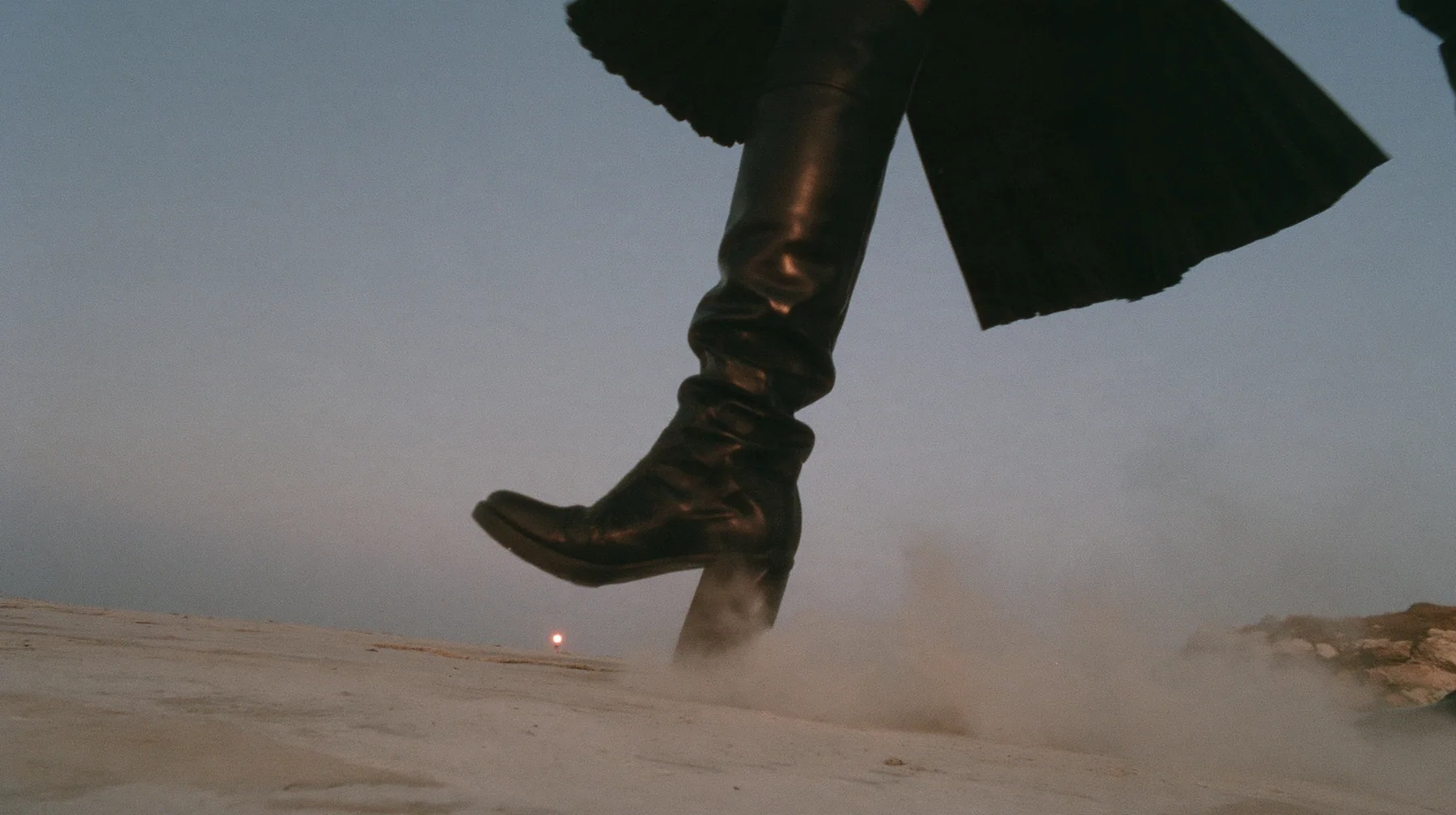 low close-up of a black knee boot stamping down on pale concrete, a puff of dust, a pleated black skirt hem at the top of the frame, no face, pale dawn grey-white, soft even light, a sea of fog below, one small…