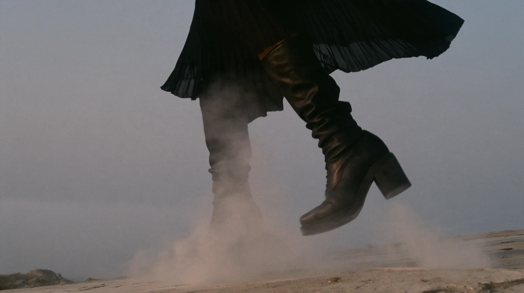 low close-up of a black knee boot stamping down on pale concrete, a puff of dust, a pleated black skirt hem at the top of the frame, no face, pale dawn grey-white, soft even light, a sea of fog below, one small…