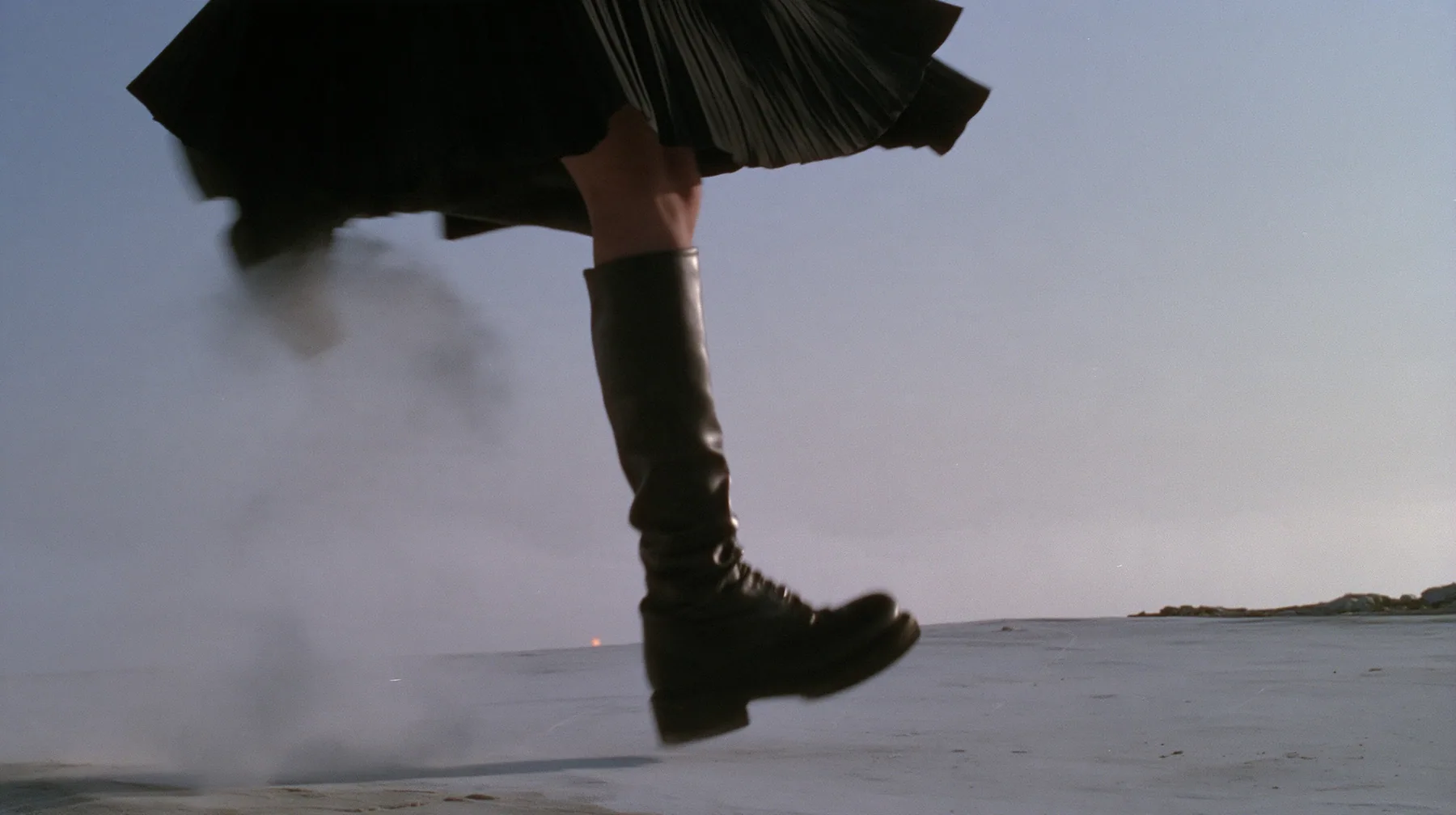 low close-up of a black knee boot stamping down on pale concrete, a puff of dust, a pleated black skirt hem at the top of the frame, no face, pale dawn grey-white, soft even light, a sea of fog below, one small…