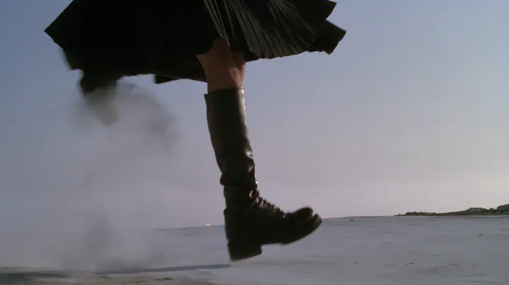 low close-up of a black knee boot stamping down on pale concrete, a puff of dust, a pleated black skirt hem at the top of the frame, no face, pale dawn grey-white, soft even light, a sea of fog below, one small…