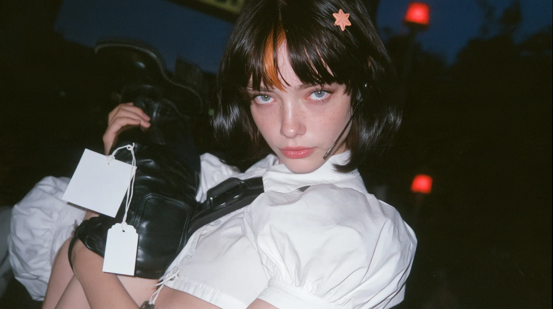 medium close-up of a young Caucasian American woman with pale skin and light freckles, a glossy black blunt jaw-length bob with heavy bangs and one clay-orange streak through the bangs, a small flat clay-orange…
