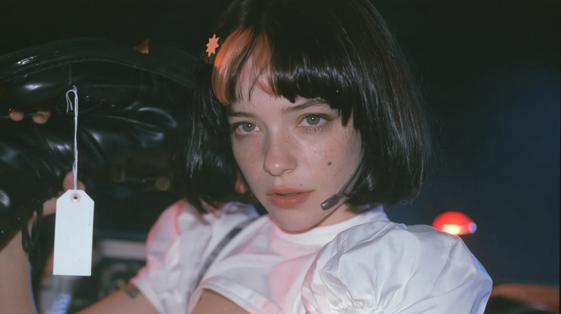 medium close-up of a young Caucasian American woman with pale skin and light freckles, a glossy black blunt jaw-length bob with heavy bangs and one clay-orange streak through the bangs, a small flat clay-orange…