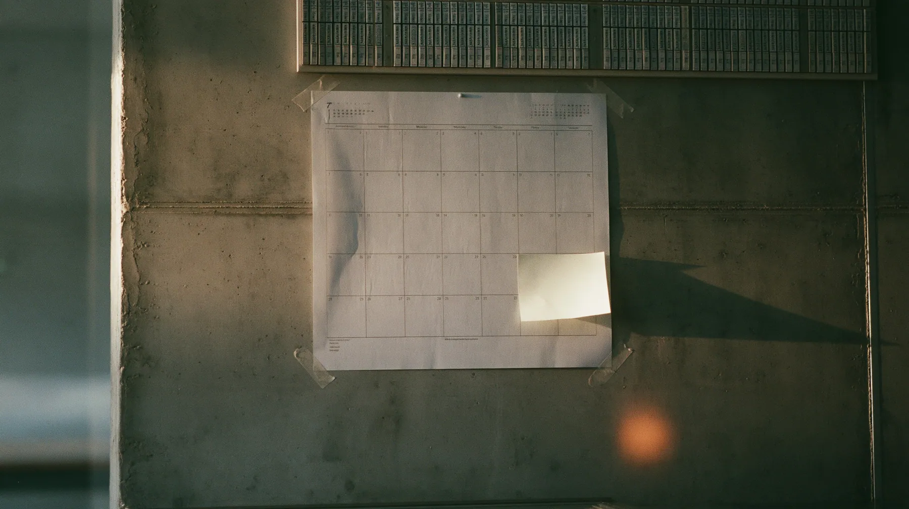 a paper wall calendar with an empty month grid taped to raw concrete below a giant split-flap board, one blank off-white sticky note stuck to it, pale dawn light raking across, pale dawn grey-white, soft even light,…