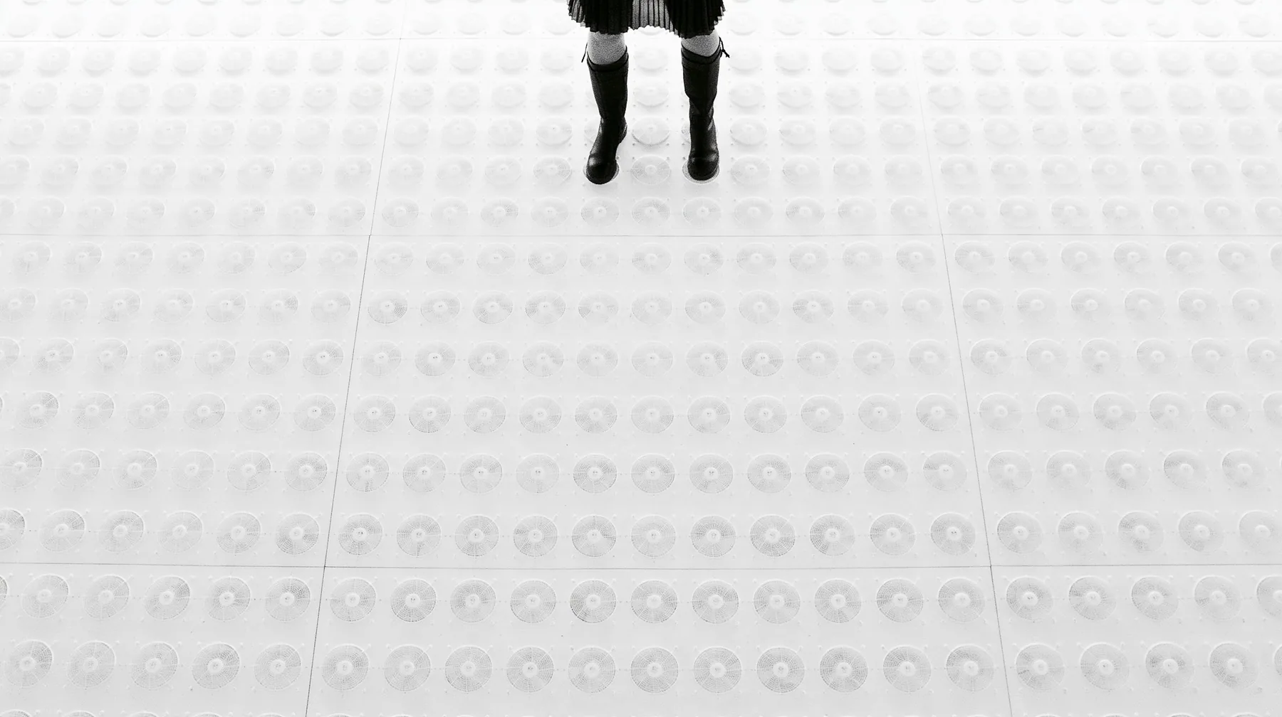 straight-down aerial of a vast datacenter roof with hundreds of identical round cooling fans in a grid like a city, white haze, the black pleated skirt hem and two black knee boots at the top edge of the frame,…