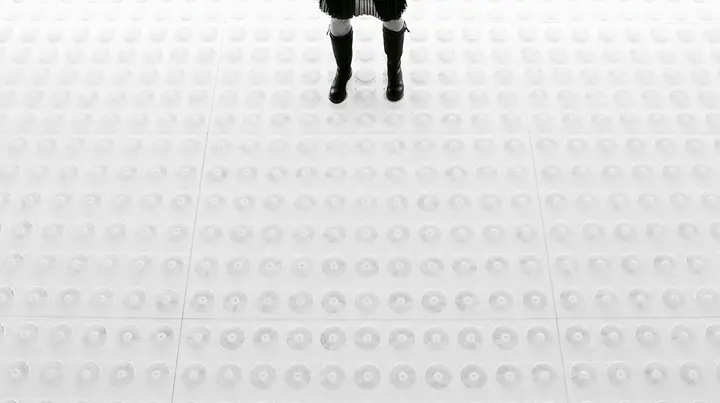 straight-down aerial of a vast datacenter roof with hundreds of identical round cooling fans in a grid like a city, white haze, the black pleated skirt hem and two black knee boots at the top edge of the frame,…
