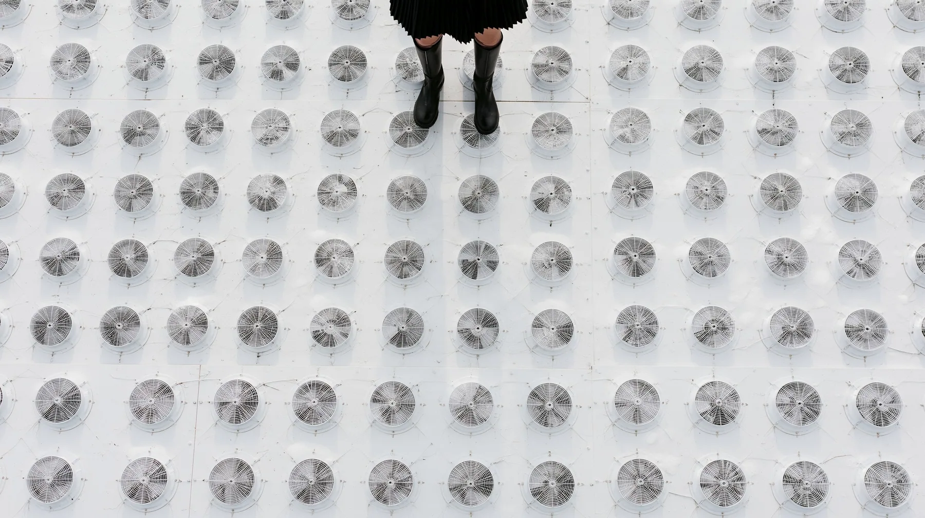 straight-down aerial of a vast datacenter roof with hundreds of identical round cooling fans in a grid like a city, white haze, the black pleated skirt hem and two black knee boots at the top edge of the frame,…