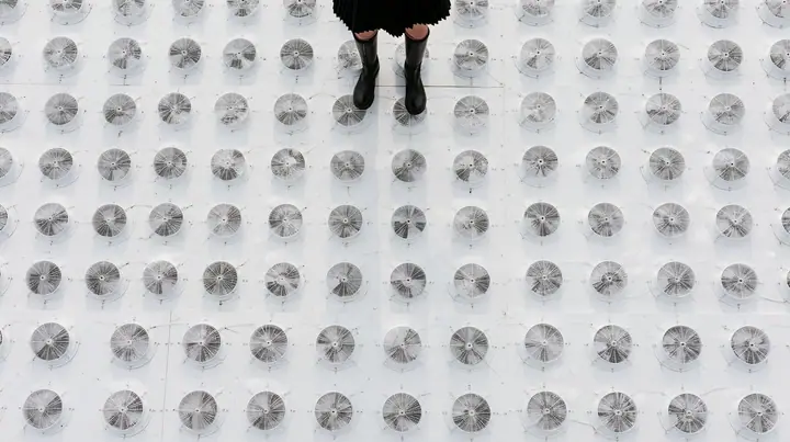 straight-down aerial of a vast datacenter roof with hundreds of identical round cooling fans in a grid like a city, white haze, the black pleated skirt hem and two black knee boots at the top edge of the frame,…