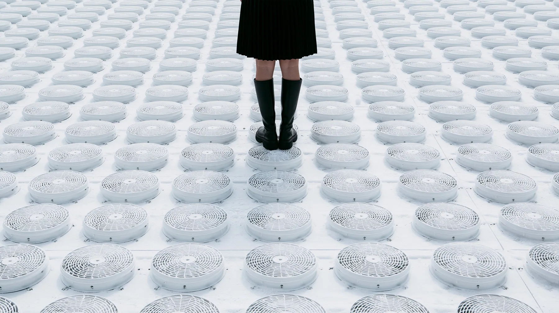 straight-down aerial of a vast datacenter roof with hundreds of identical round cooling fans in a grid like a city, white haze, the black pleated skirt hem and two black knee boots at the top edge of the frame,…