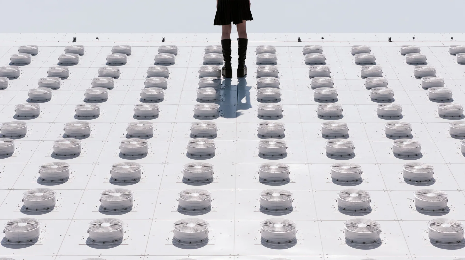 straight-down aerial of a vast datacenter roof with hundreds of identical round cooling fans in a grid like a city, white haze, the black pleated skirt hem and two black knee boots at the top edge of the frame,…