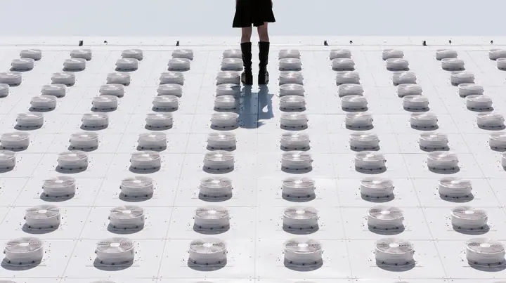 straight-down aerial of a vast datacenter roof with hundreds of identical round cooling fans in a grid like a city, white haze, the black pleated skirt hem and two black knee boots at the top edge of the frame,…
