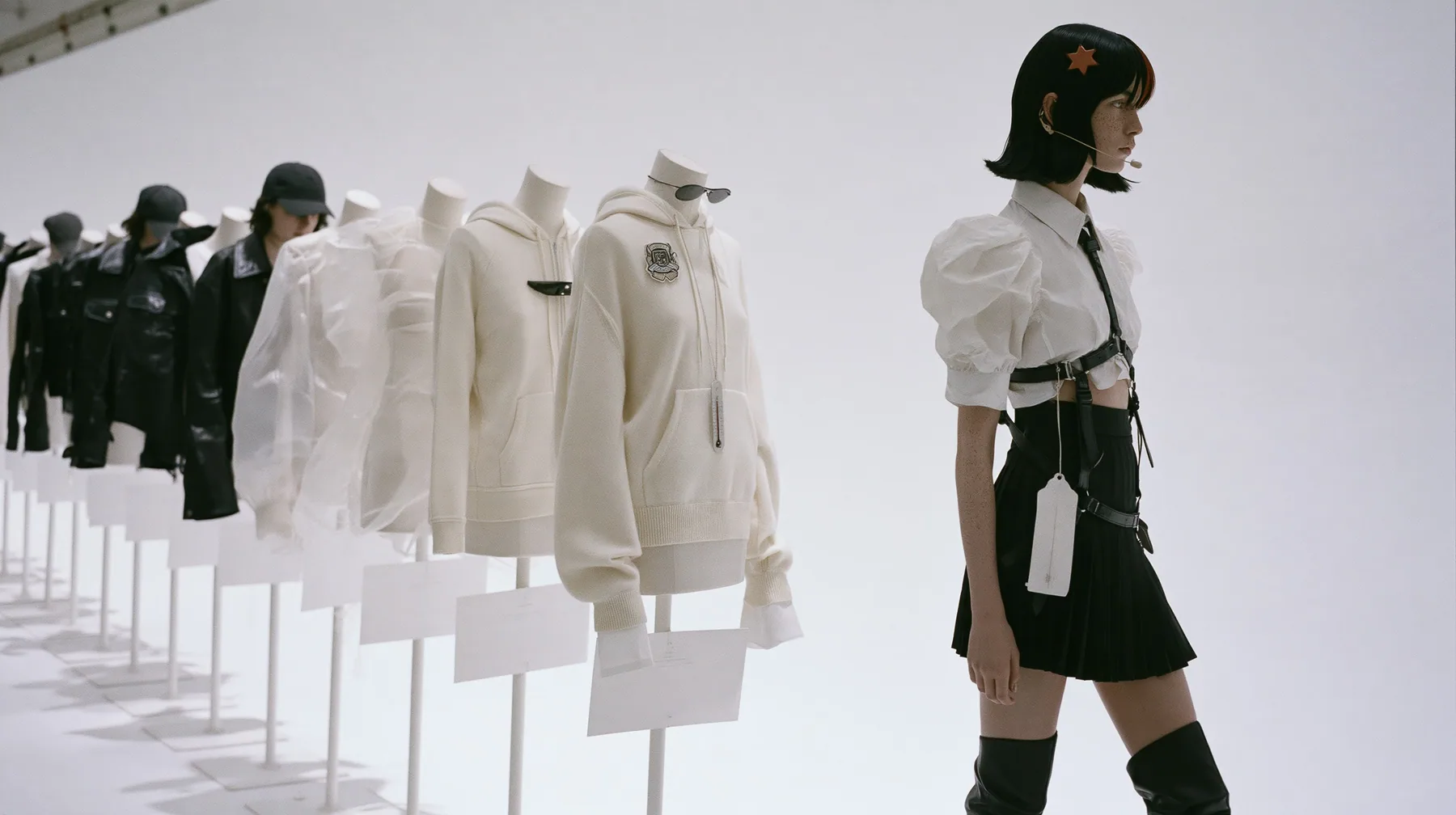wide shot of eleven headless dress forms lined along an endless white catwalk, dressed in a washed black dad cap, a black leather jacket, a sheer organza shell, a pale cashmere hoodie with a badge, narrow sunglasses,…