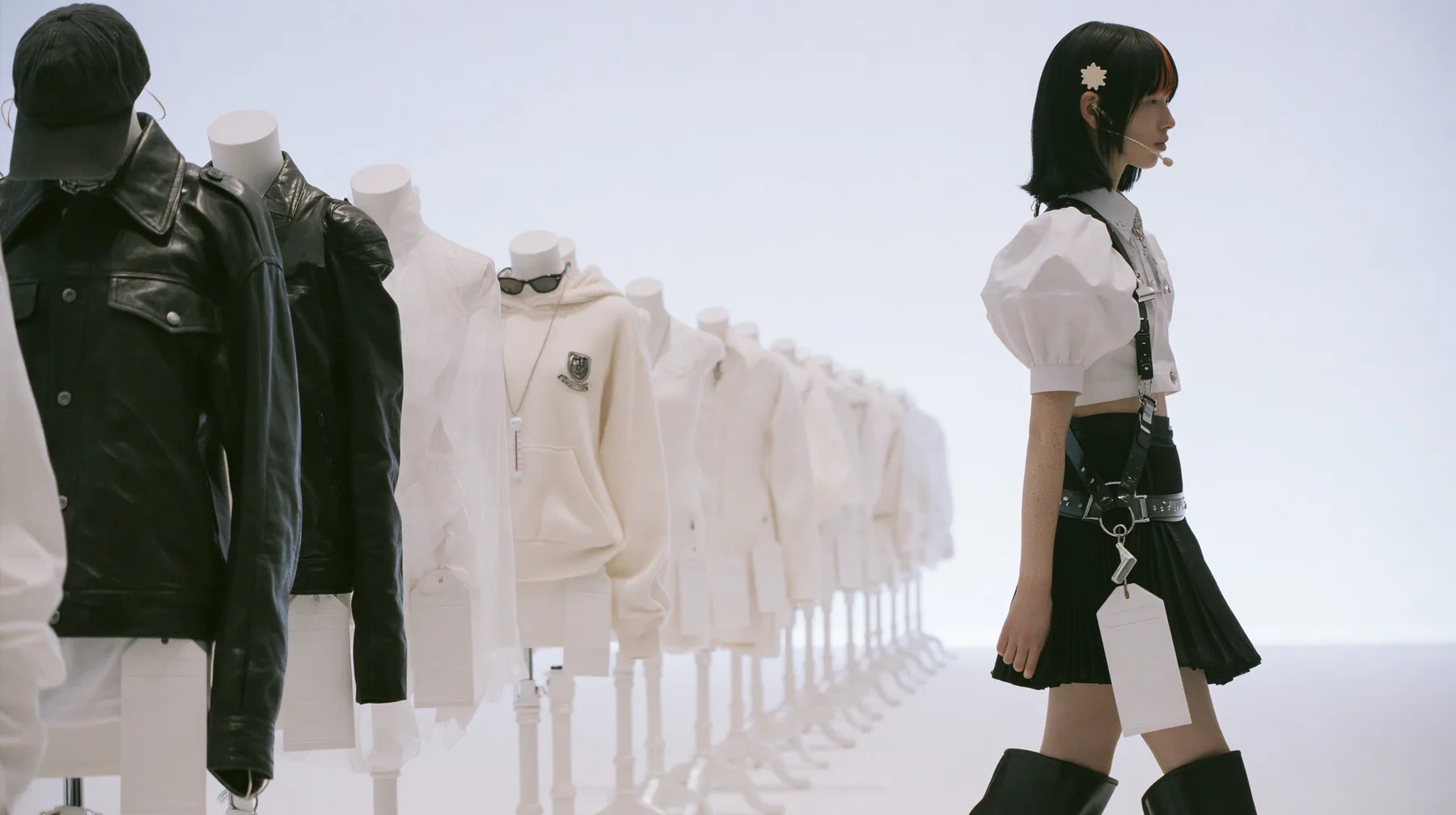 wide shot of eleven headless dress forms lined along an endless white catwalk, dressed in a washed black dad cap, a black leather jacket, a sheer organza shell, a pale cashmere hoodie with a badge, narrow sunglasses,…