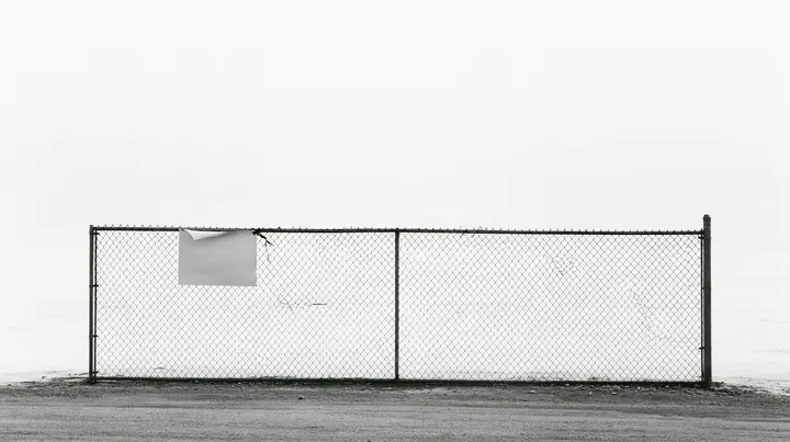 wide shot of a chain-link construction fence at the edge of a vast empty desert site, one blank laminated notice cable-tied to it and flapping, high-key white sky, no people, high-key white paper light, almost no…