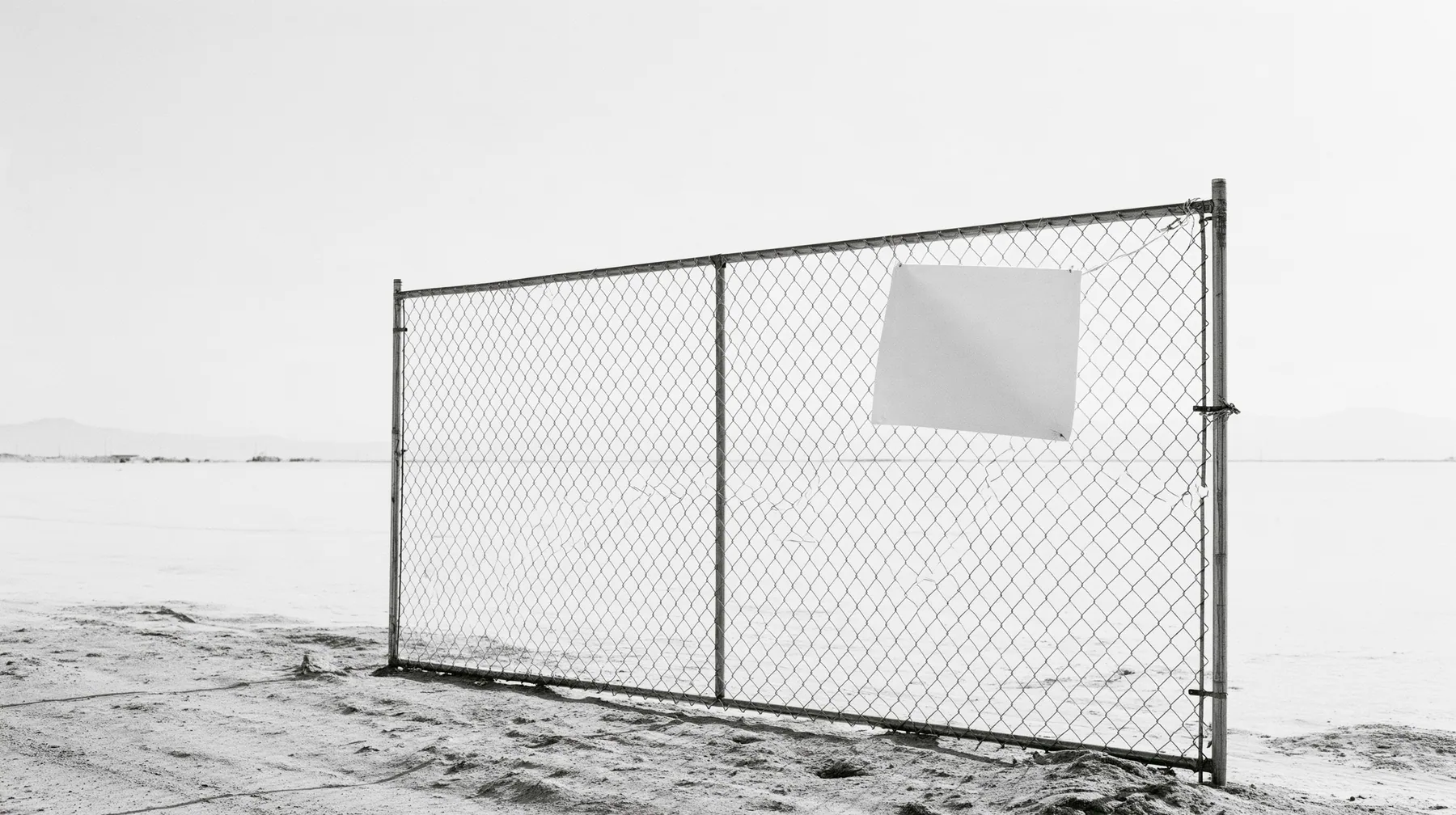 wide shot of a chain-link construction fence at the edge of a vast empty desert site, one blank laminated notice cable-tied to it and flapping, high-key white sky, no people, high-key white paper light, almost no…