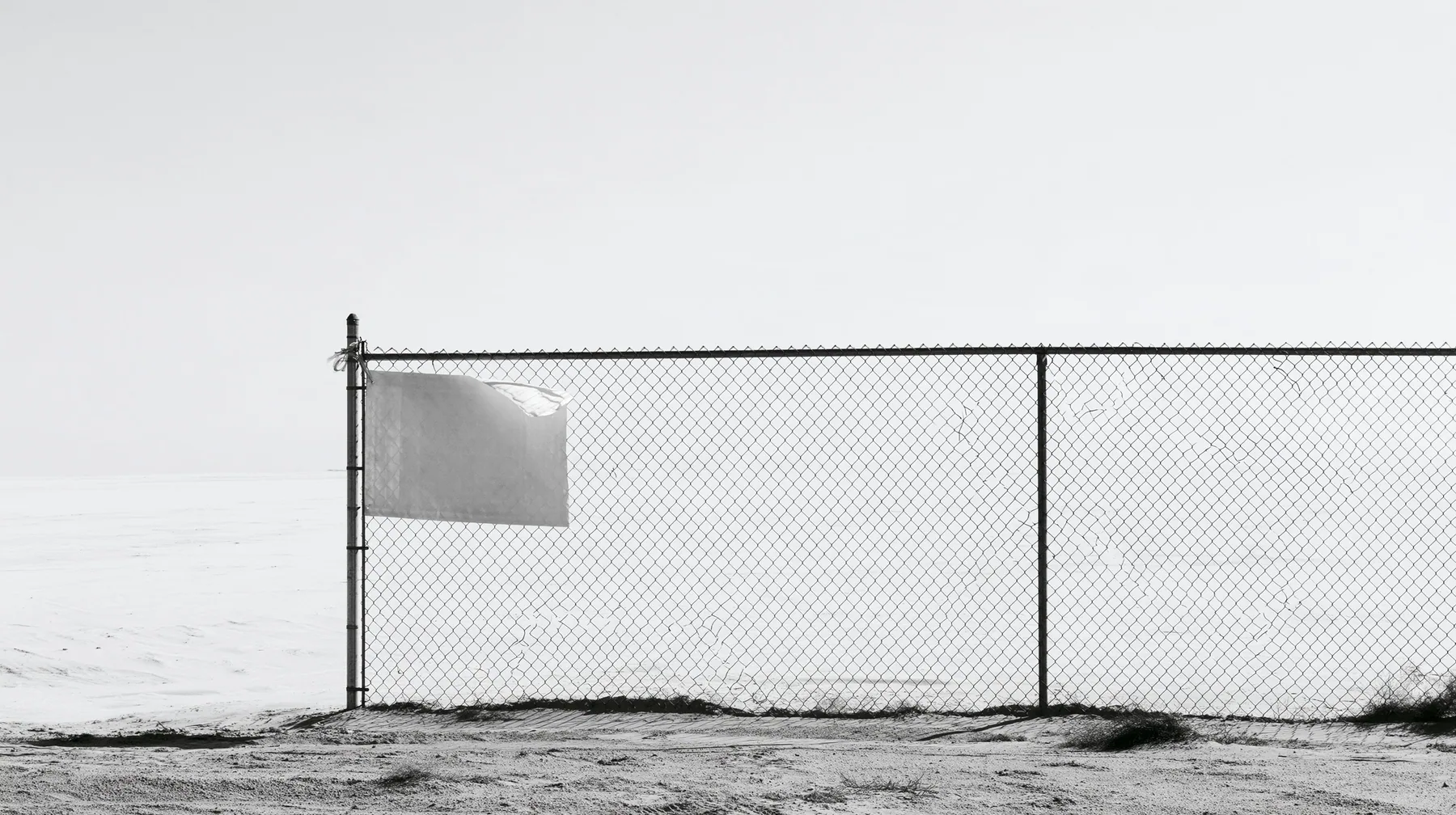 wide shot of a chain-link construction fence at the edge of a vast empty desert site, one blank laminated notice cable-tied to it and flapping, high-key white sky, no people, high-key white paper light, almost no…