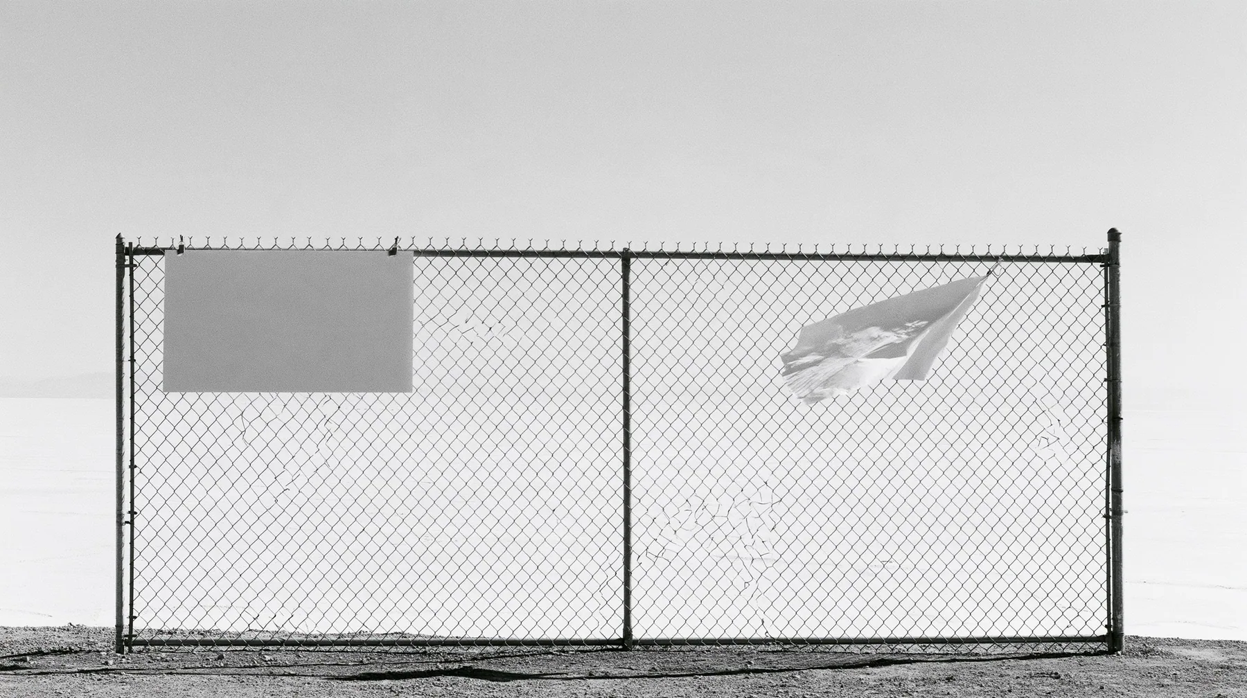 wide shot of a chain-link construction fence at the edge of a vast empty desert site, one blank laminated notice cable-tied to it and flapping, high-key white sky, no people, high-key white paper light, almost no…