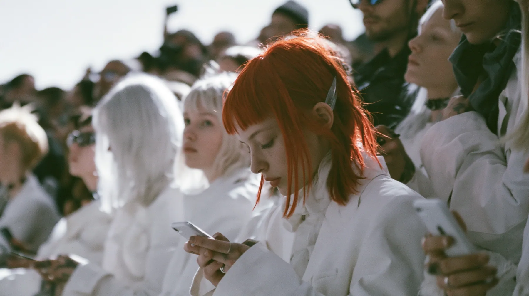 wide shot of a fashion show front row in high-key white light, the guests standing up and putting their phones away, faces cropped at the top of the frame, high-key white paper, soft shadowless light, monochrome…