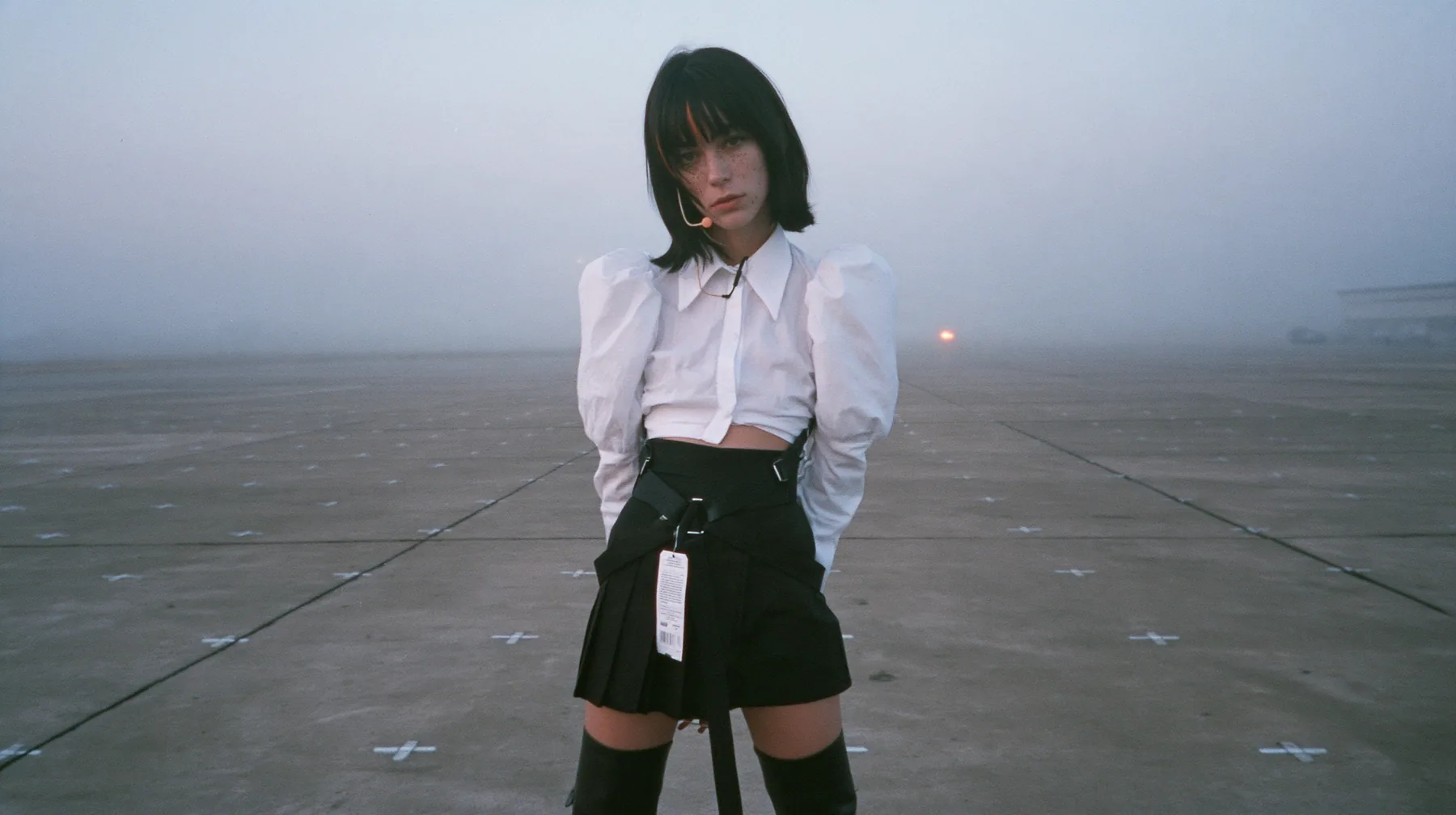 extreme wide shot of a vast empty grey concrete hall with a perfect grid of small painted floor marks receding to the horizon, a young Caucasian American woman with pale skin and light freckles, a glossy black blunt…