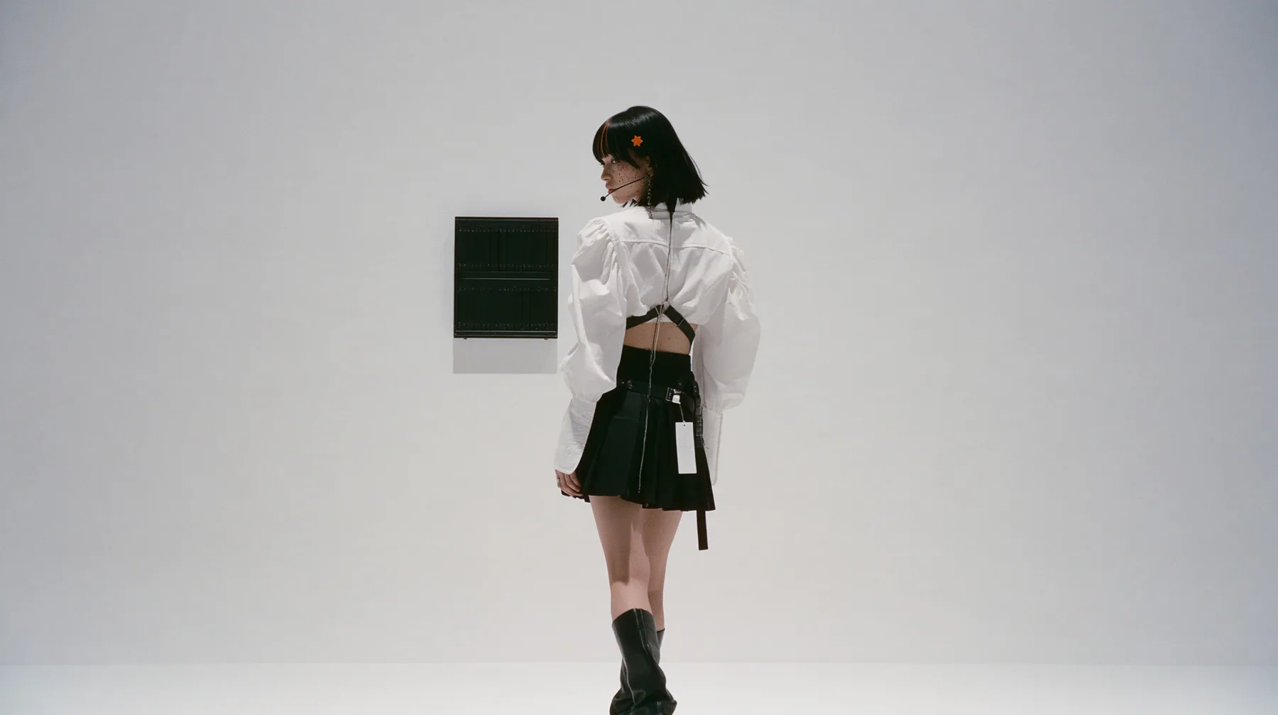wide shot of a small black split-flap board with blank flaps alone on a seamless white paper wall, a young Caucasian American woman with pale skin and light freckles, a glossy black blunt jaw-length bob with heavy…